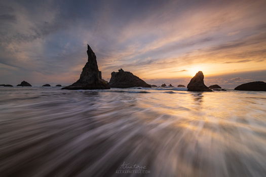 Silhouetted sea stacks rising from shallow ocean waters at sunset with soft wave motion blur.