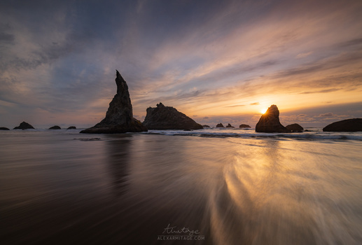 Dramatic sea stacks silhouetted against a golden sunset sky with smooth water in the foreground.
