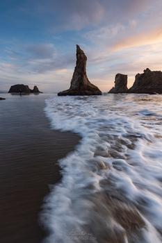 Dramatic rocky sea stacks rising from foaming waves at sunset with golden-hour light illuminating the cloudy sky.