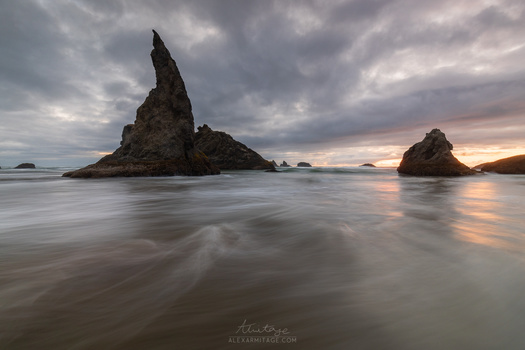 Tall rocky sea stack silhouetted against dramatic cloudy sky at sunset, with smooth water in foreground.
