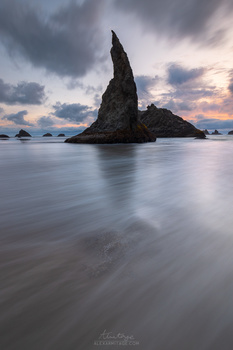 Tall rock formation rising from calm ocean waters at twilight, surrounded by smaller sea stacks and dramatic cloudy sky.