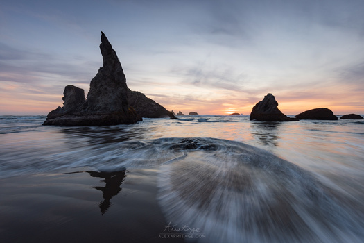 Dramatic rocky sea stacks silhouetted against a twilight sky with waves washing across the foreground beach.