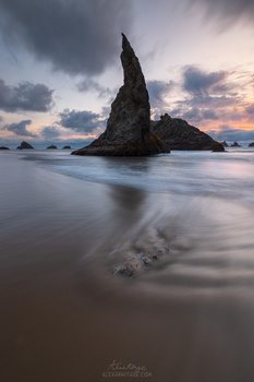 Tall rock formation rising from shallow ocean water at twilight, with smooth waves flowing across sandy beach.