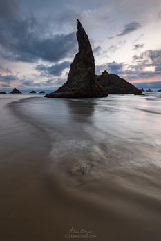 Tall sea stack rising from shallow water at dusk with dramatic cloudy sky.