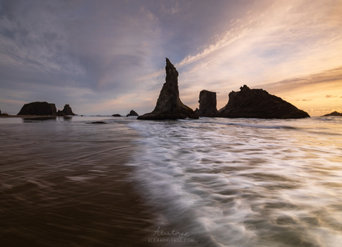 Dramatic rocky sea stacks silhouetted against a golden sunset sky with waves washing across the foreground beach.