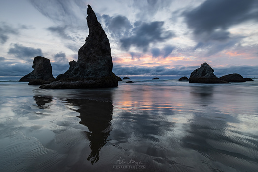 Tall rock formation reflected in wet sand at twilight, with smaller sea stacks and cloudy sky.