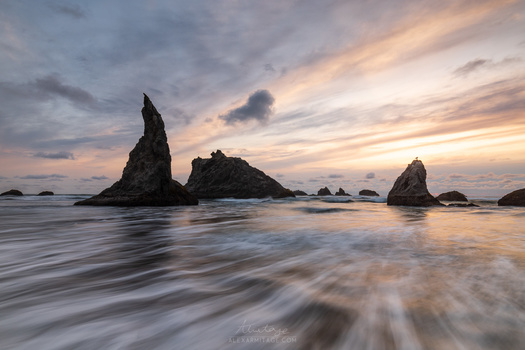 Dramatic rock formations rising from the ocean at sunset, with smooth water and golden sky.