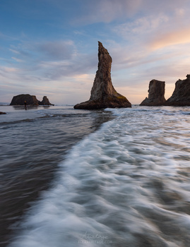 Tall sea stack rising from shallow water with waves washing across the beach at golden hour.