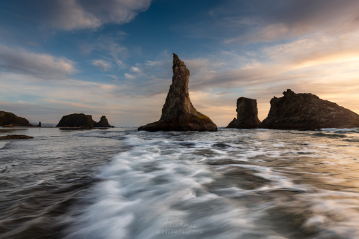 Dramatic sea stacks rising from the ocean during golden hour with smooth water and dynamic clouds.