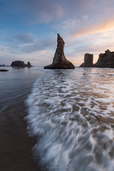 Dramatic rock formations rise from the ocean during sunset with flowing waves in the foreground.