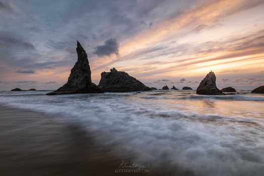 Dramatic coastal rock formations silhouetted against a golden and purple sunset sky with waves washing across the beach.