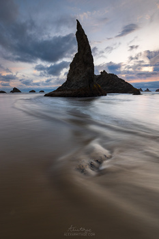 Dark rocky sea stack rising from shallow water at dusk, with soft wave motion in foreground.
