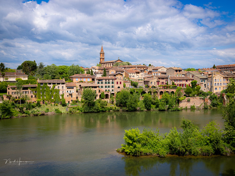 Medieval hilltop town with terracotta buildings and church tower reflected in river below.
