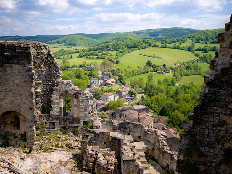 Stone castle ruins frame a verdant valley landscape with scattered villages and forested hills under partly cloudy skies.