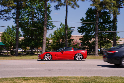 Bright red sports car photographed in profile on a suburban street lined with tall trees.