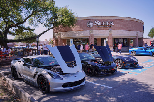 Three Corvettes with open hoods displayed outdoors at a car show near a brick building.
