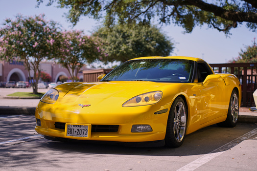 Bright yellow sports car parked in sunny outdoor lot with flowering trees and architecture visible in background.