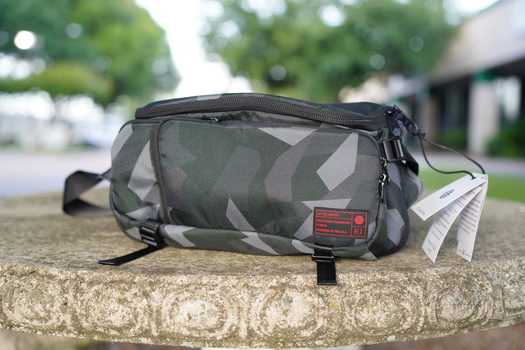Gray camouflage camera sling bag with shoulder strap positioned on textured concrete surface.