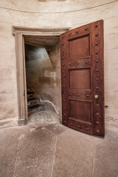 Ornate wooden door with decorative studs open to reveal stone interior chamber with dramatic sunlight streaming through window.