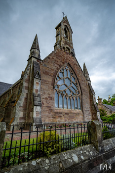 Gothic stone chapel with clock tower beneath overcast sky, framed by iron fence.
