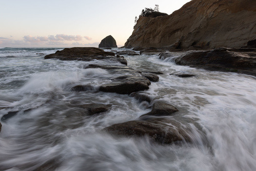 Waves crash over dark rocks on a rocky beach at dusk, with a sea stack and forested headland visible in the background.