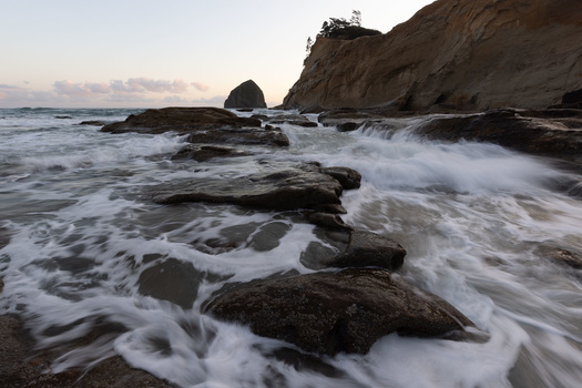 Ocean waves crashing over dark rocks at a rugged coastline during golden hour.