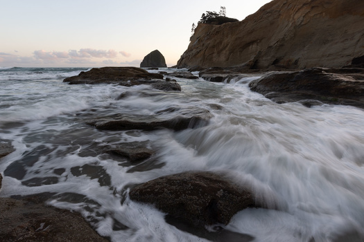 Waves crash over dark rocks on a sandy beach at dusk, with a steep cliff and isolated sea stack in the background.