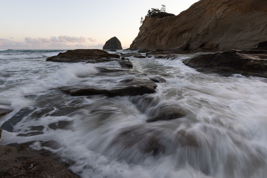 Ocean waves crash over dark rocks at a rugged coastline during twilight, with a cliff and sea stack visible in the background.