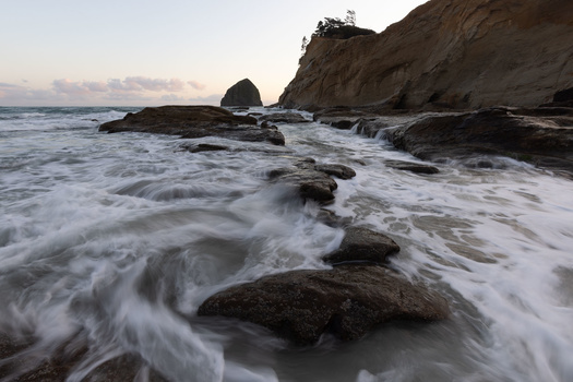 Waves crash over dark rocks on a beach at dusk, with a sea stack and forested cliff in the background.