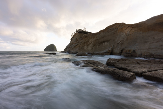 Rocky coastal shoreline with sea stack and crashing waves at dusk.
