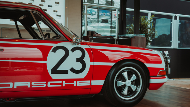 Red and white Porsche race car with number 23 displayed on the door, shot in profile in a modern showroom setting.
