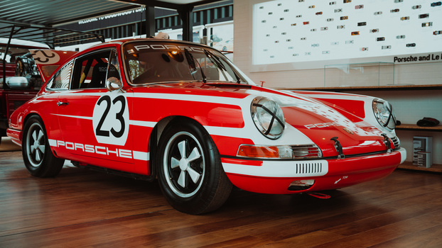 Red and white Porsche 911 race car with number 23 displayed in a showroom with wooden flooring.