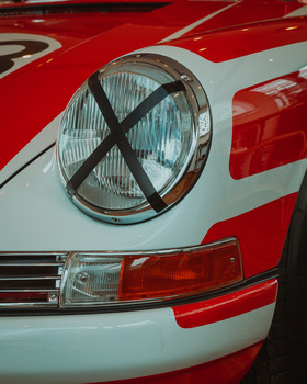 Close-up of a classic car's headlight with a black X taped across the glass, set against red and white bodywork.