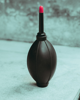 Black teardrop-shaped cosmetic applicator with a bright pink-red lipstick tip against a weathered turquoise background.