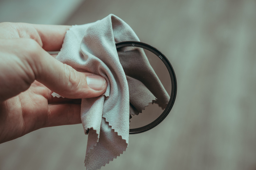 Hand holding a circular camera filter while wiping it with a light beige microfiber cloth.