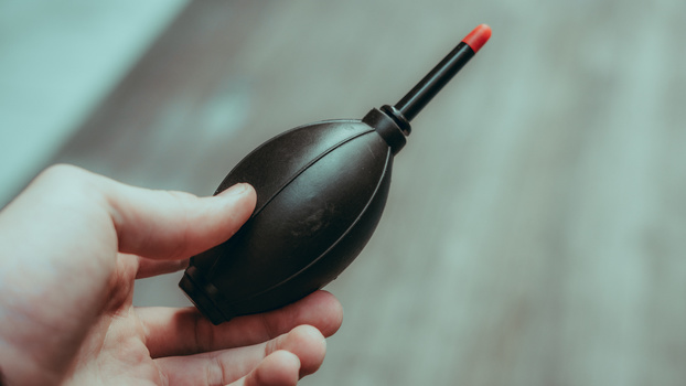 Hand holding a black air blower bulb with a red rubber tip against a blurred background.