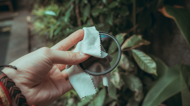 Hand holding a magnifying glass over a white fabric swatch against blurred green foliage background.