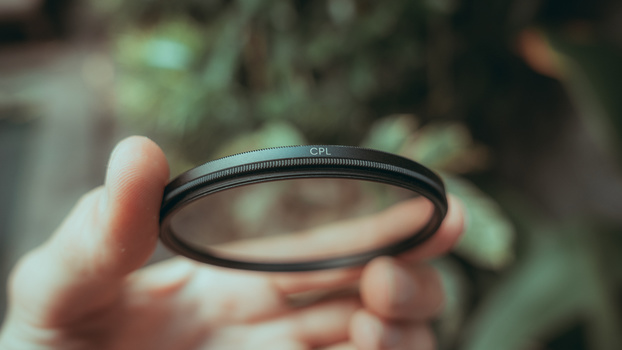 A CPL (circular polarizing) filter held in hand against a blurred outdoor background.