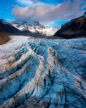 Massive glacier with deep blue crevasses stretches across a valley floor beneath snow-capped peaks.
