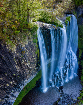 Waterfall cascading down dark basalt cliffs surrounded by moss-covered rocks and forest canopy.