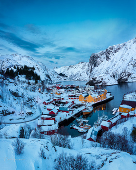 Snow-covered harbor village nestled between steep mountains at twilight.