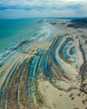 Aerial view of curved tidal patterns and sandbars creating concentric rings along a turquoise coastline.