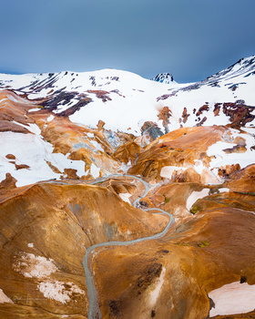 Snow-capped mountain peak above rust-colored mineral-streaked hillside with winding road.