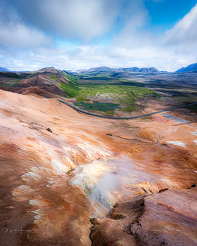 Aerial landscape showing rust-colored geothermal terrain with steaming hot springs in foreground and green valley with mountains beyond.