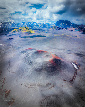 Aerial view of snow-covered volcanic landscape with colorful mineral deposits and distant snow-capped peaks.