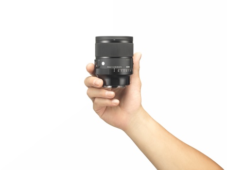 Hand holding a compact telephoto lens with focus ring and aperture markings visible.