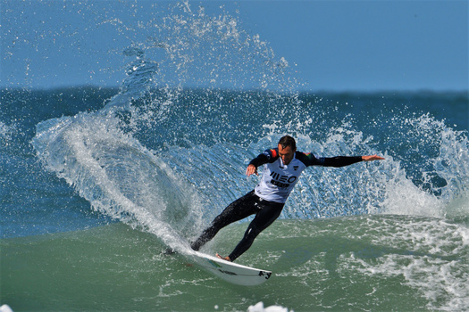 Surfer in competition attire riding a wave with dynamic spray and blue sky backdrop.