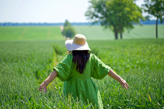 Person in green dress and white hat standing in expansive green field with arms outstretched.