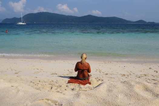Woman in rust-colored shirt sitting in meditation pose on pristine white sand beach facing turquoise water.