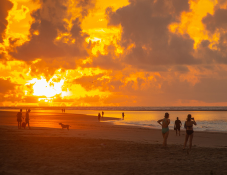 Silhouetted beachgoers wade in the ocean at sunset beneath dramatic golden clouds.